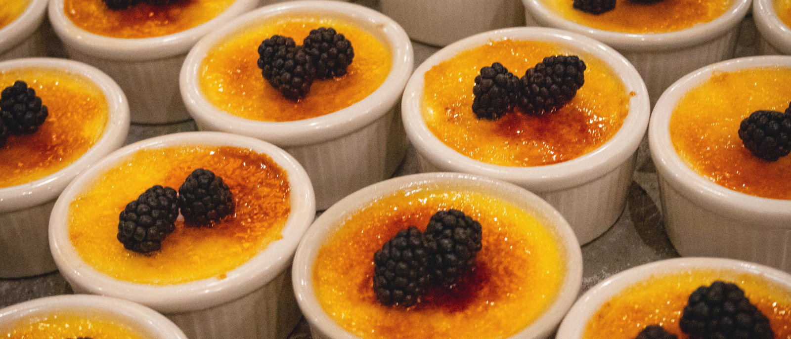 Rows of crème brûlée ramekins with caramelized sugar tops, each one finished with a single blackberry.